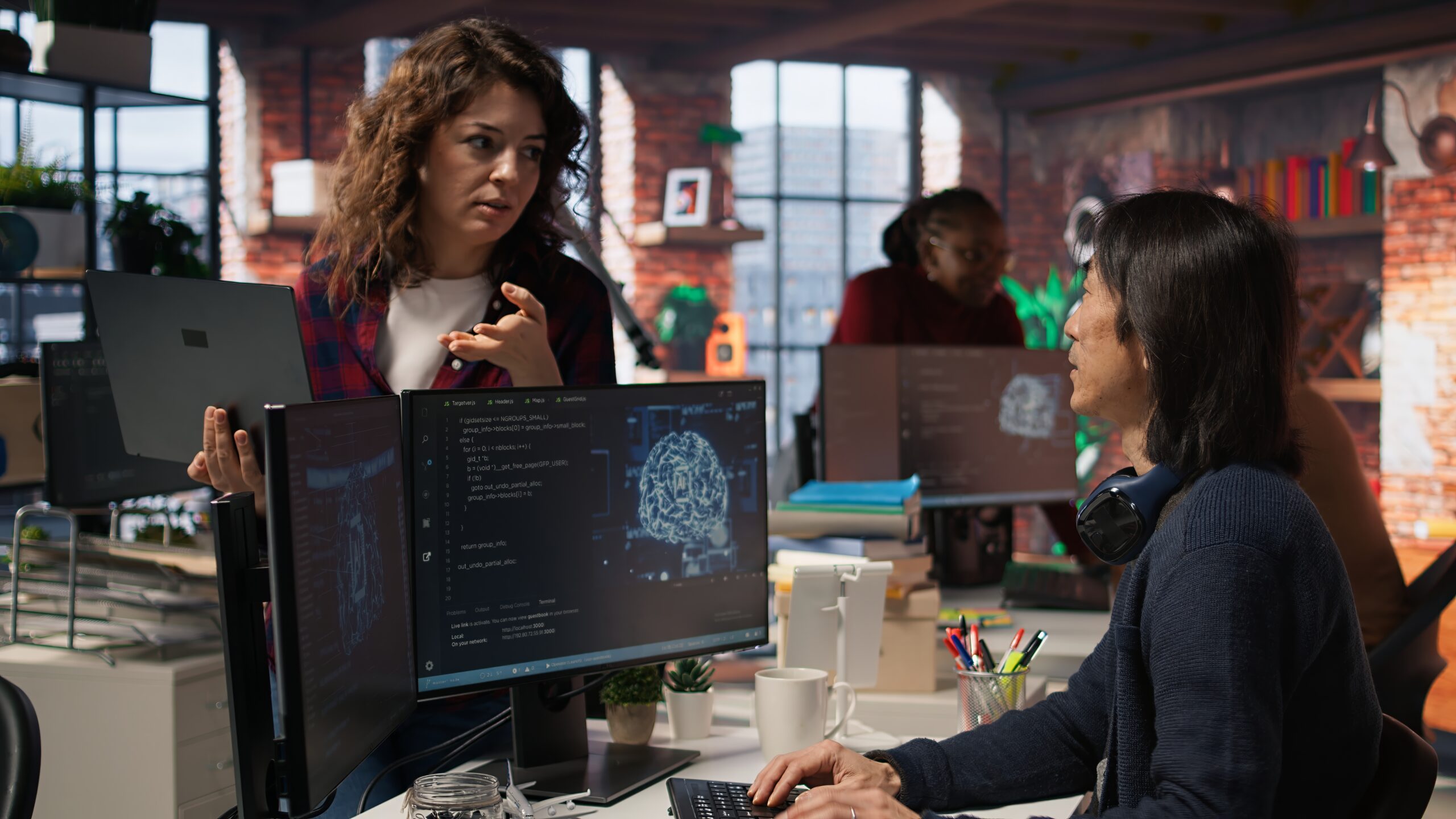 Two women collaborate at a shared desk in a modern open-office; monitors show code and a brain image on the screens.
