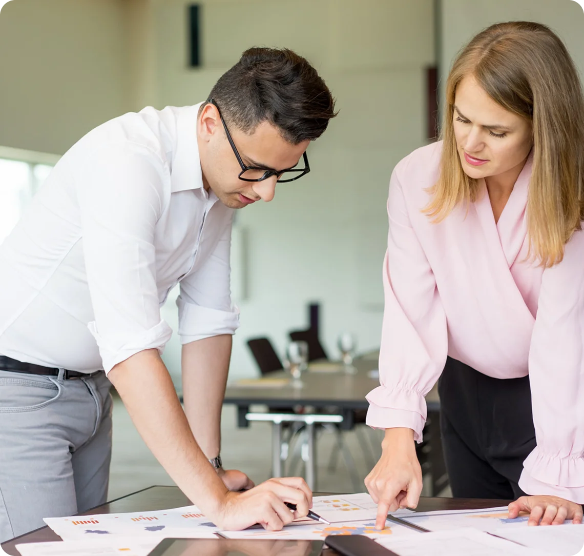 A professional man wearing glasses and a woman reviewing documents and charts together at an office desk.