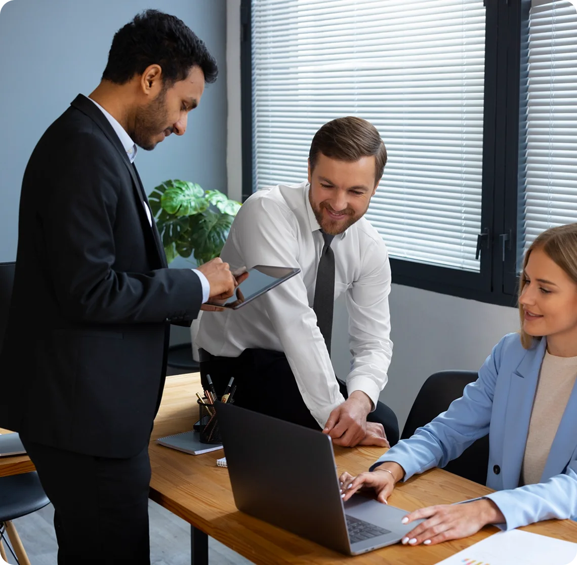 A team of three professionals collaborating in an office while using a laptop and tablet.